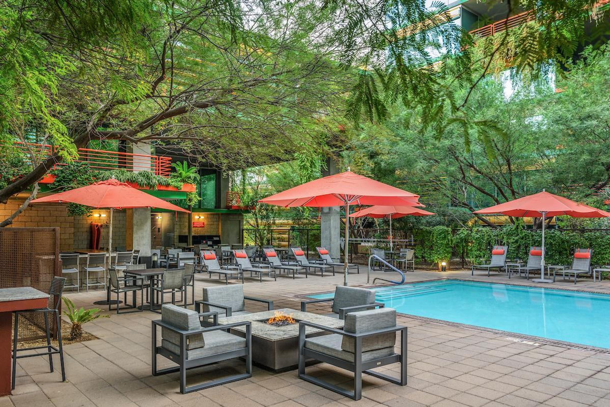Optima Sonoran Village apartments outdoor pool area with lounge chairs, red umbrellas, fire pit tables, and lush trees surrounding the space.