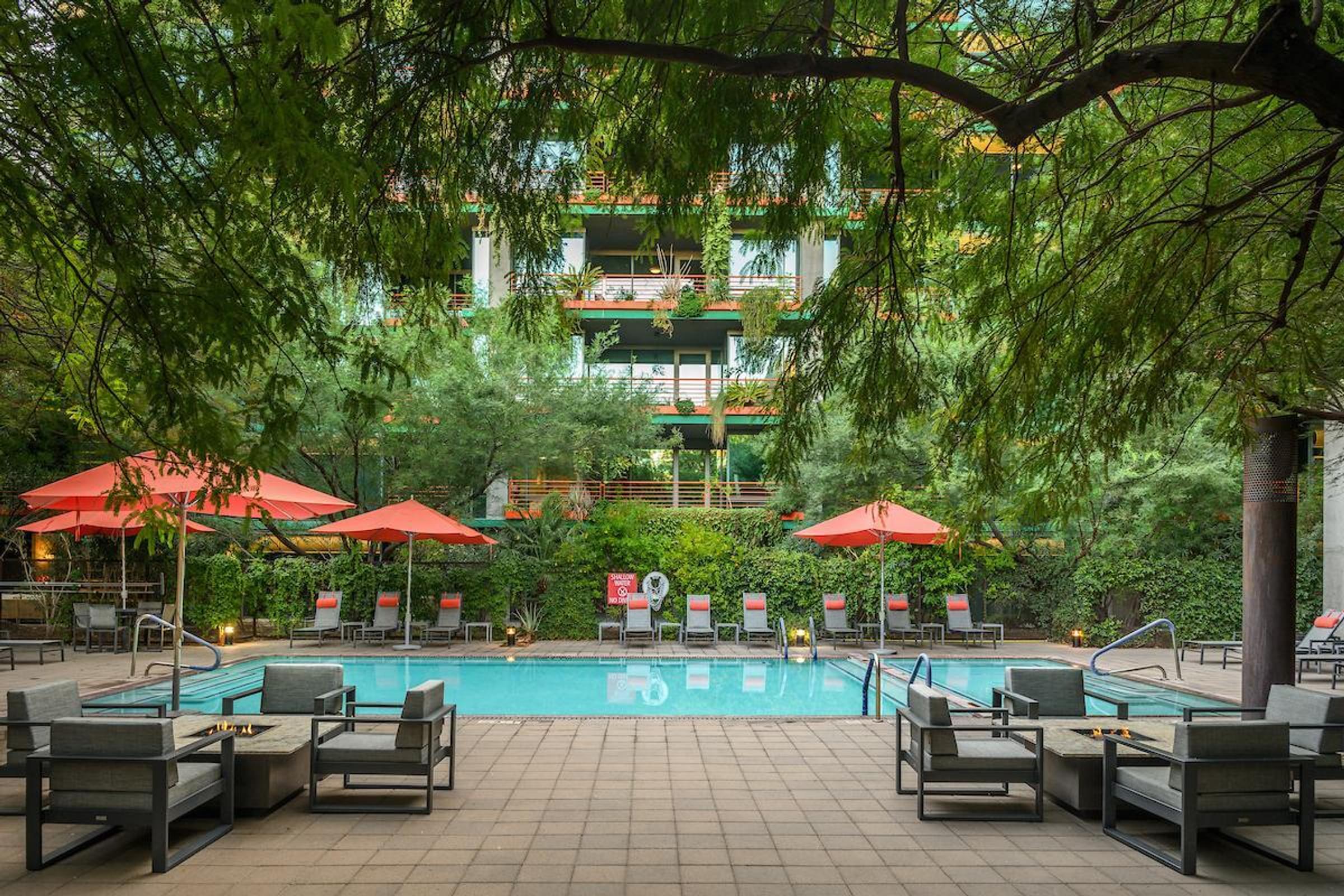 Optima Sonoran Village apartments outdoor pool area with lounge chairs, red umbrellas, patio seating, and lush greenery under a shaded tree.
