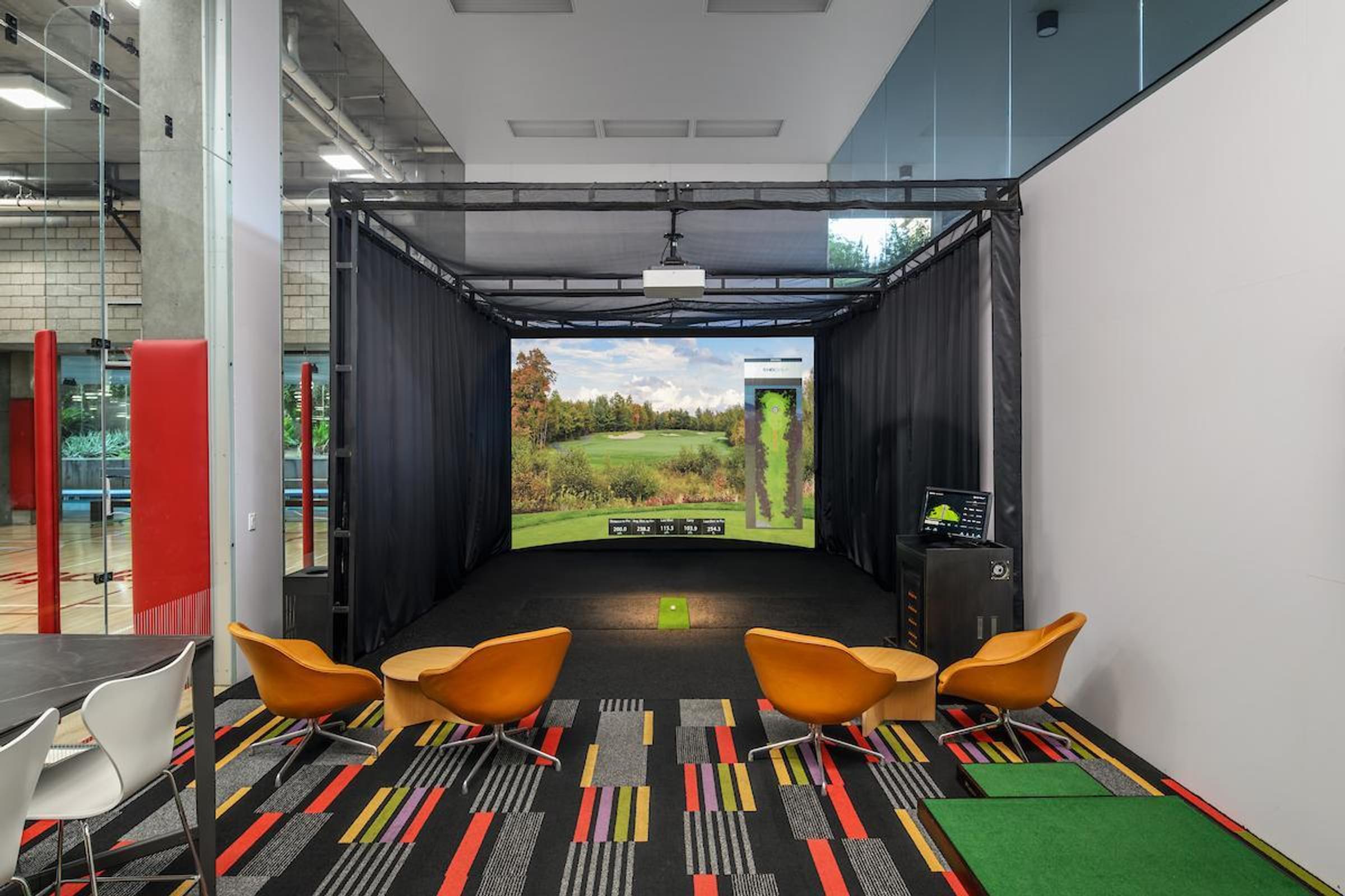 Optima Sonoran Village apartments indoor golf simulator with four yellow chairs, colorful carpet, and a large golf course screen.