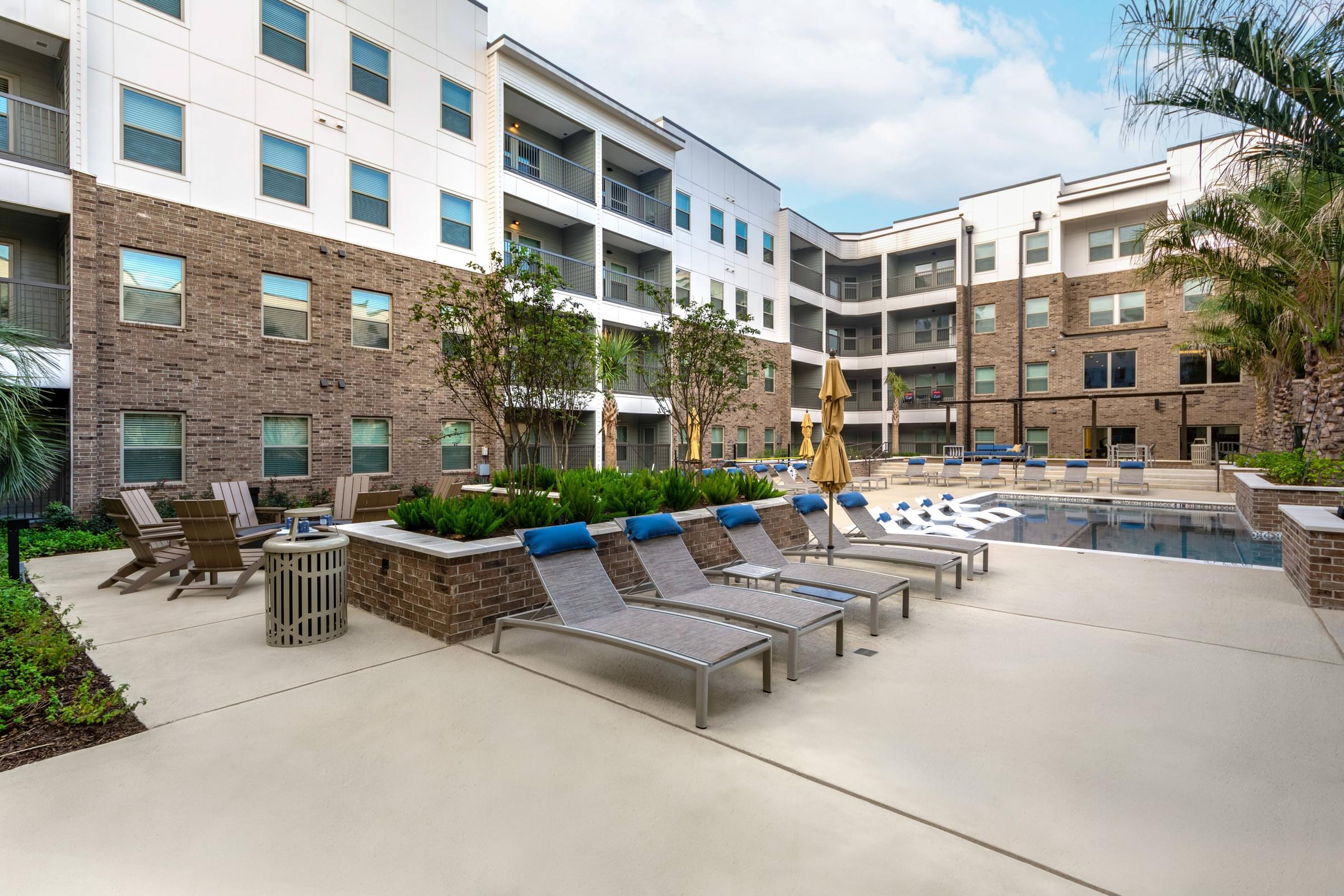 A pool surrounded by lounge chairs and umbrellas in front of apartment buildings.