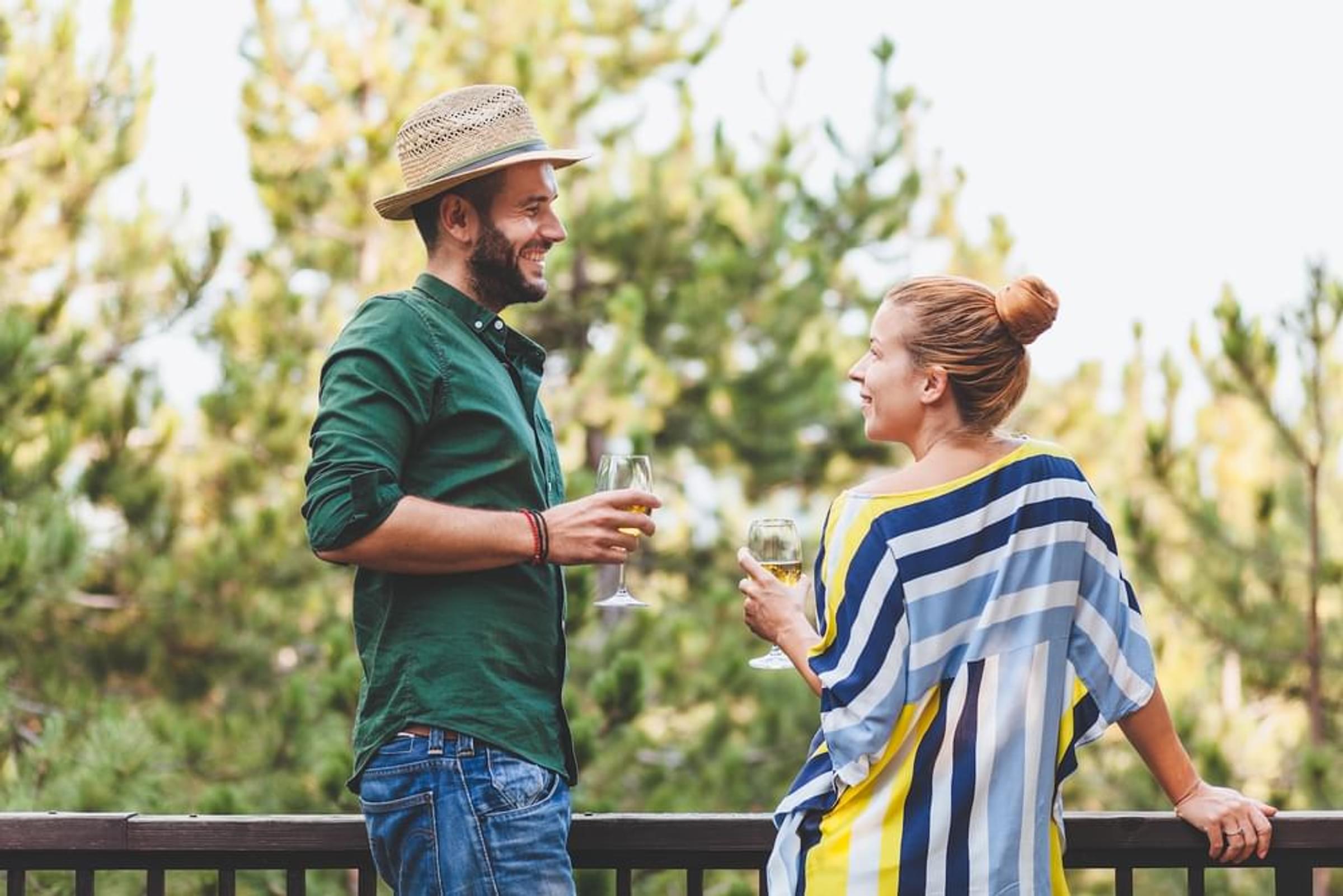 A man and a woman are standing on a balcony, both holding glasses of wine.