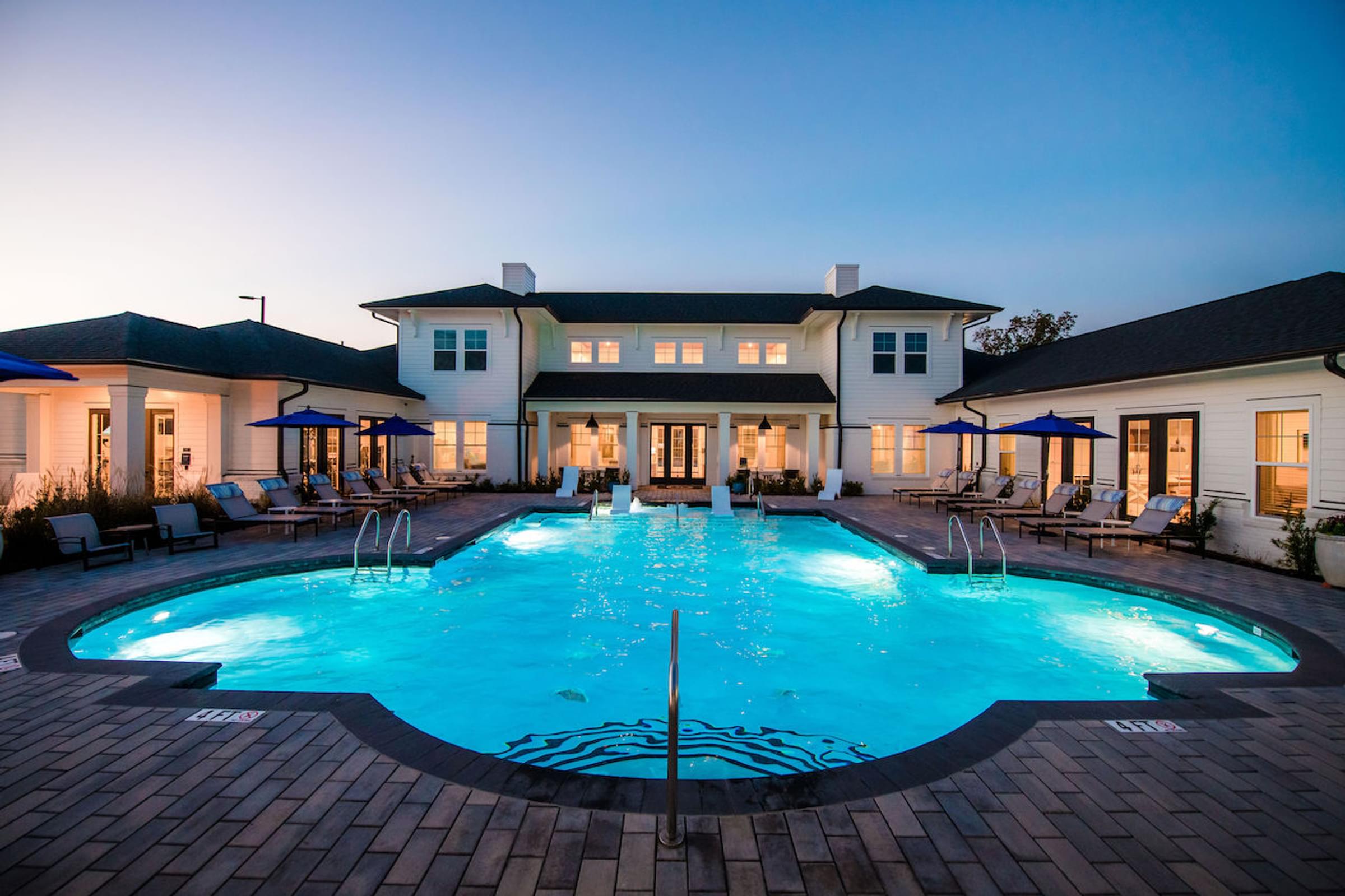a large swimming pool with lounge chairs and umbrellas in front of a large white house