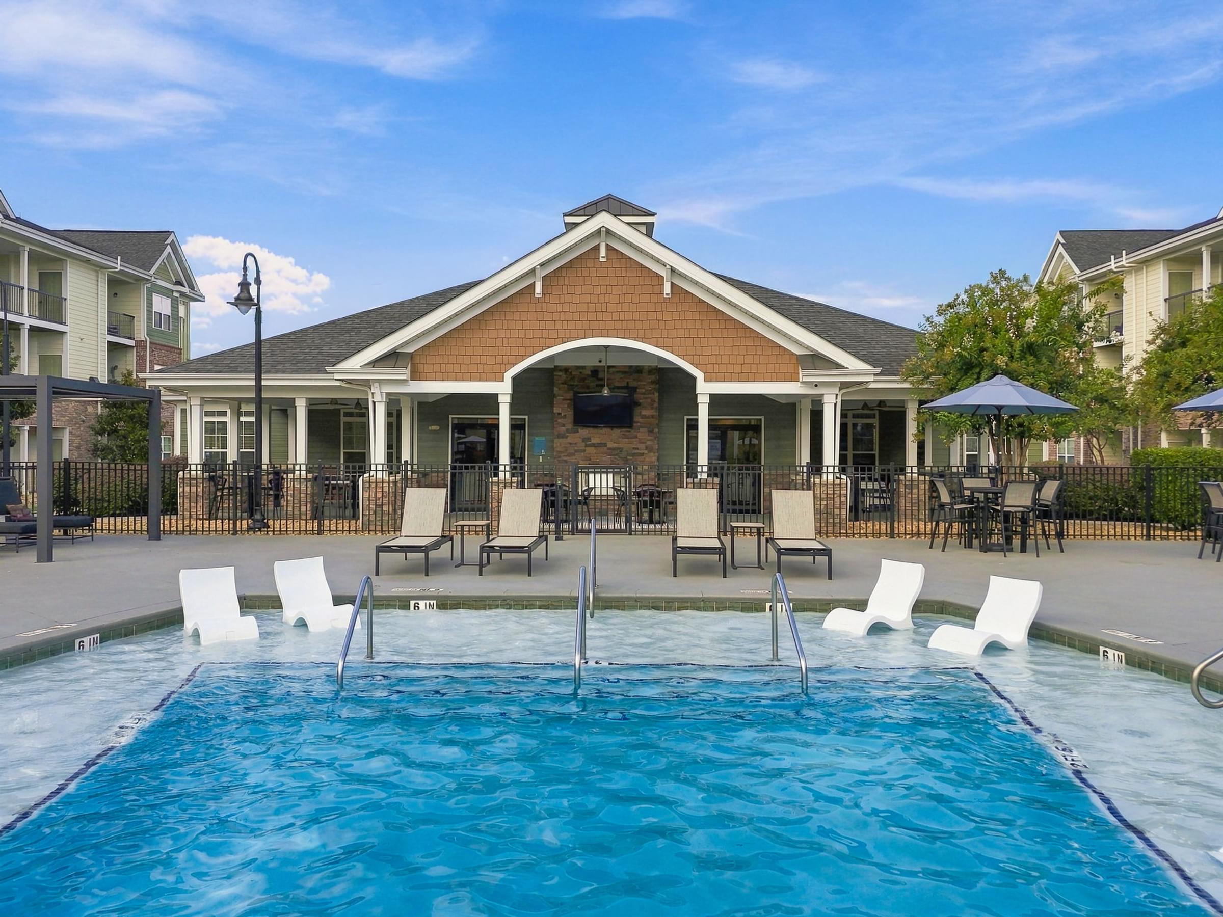 A swimming pool in front of a building with white chairs around it.