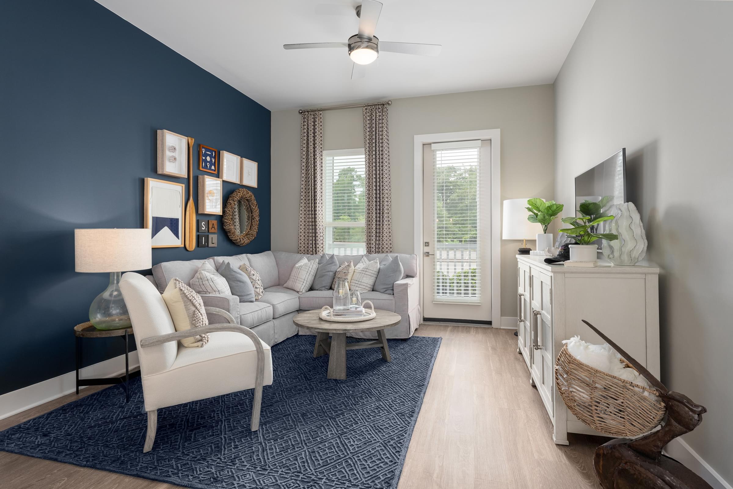 A living room with a grey sofa, a white chair, a grey coffee table, a blue rug, a ceiling fan, and a window with white curtains.
