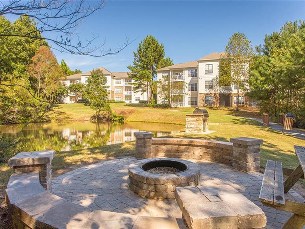 a stone fire pit and picnic table in front of a lake and an apartment building