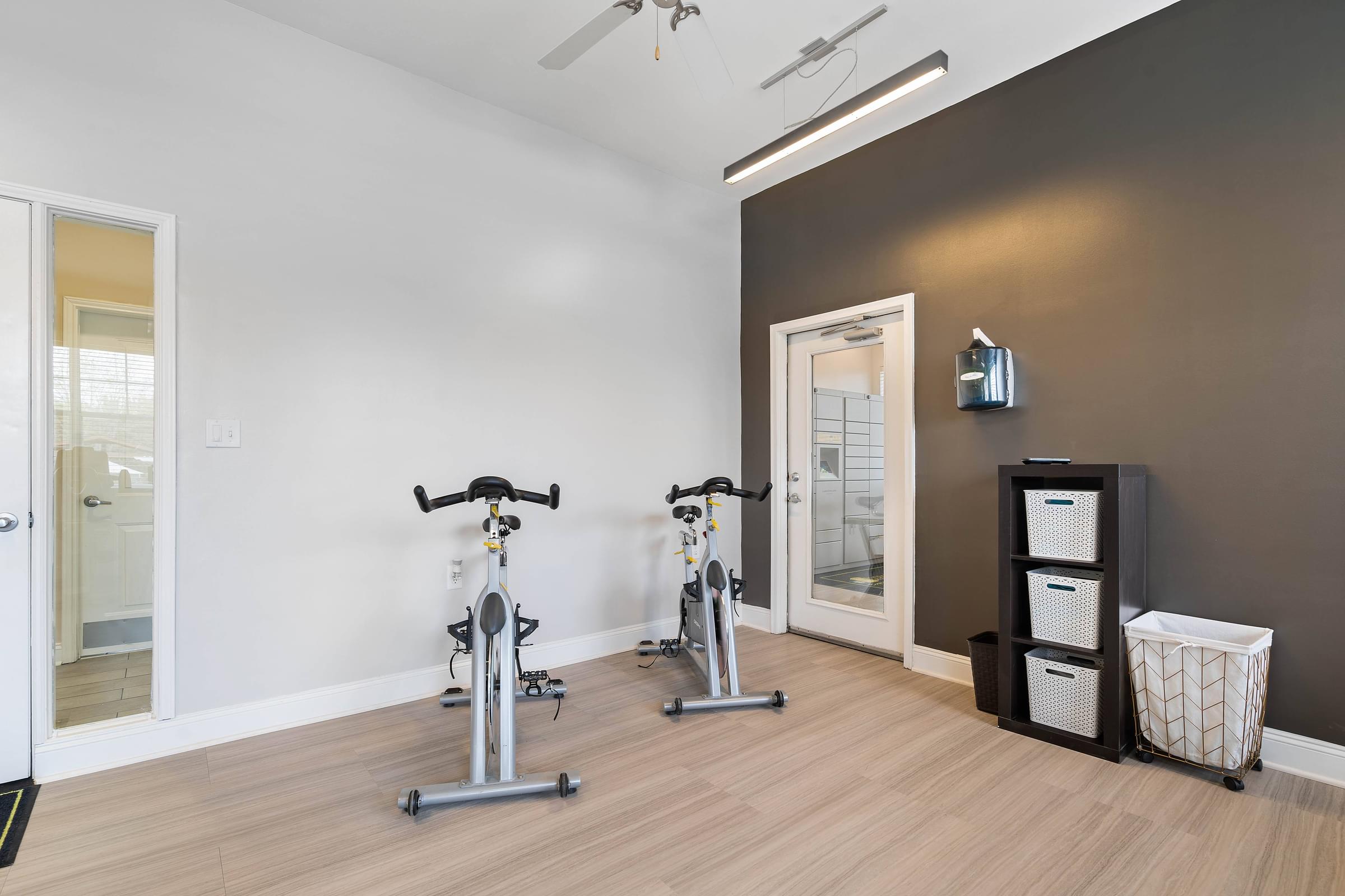 Two exercise bikes are placed in a room with a white wall and a brown wall.