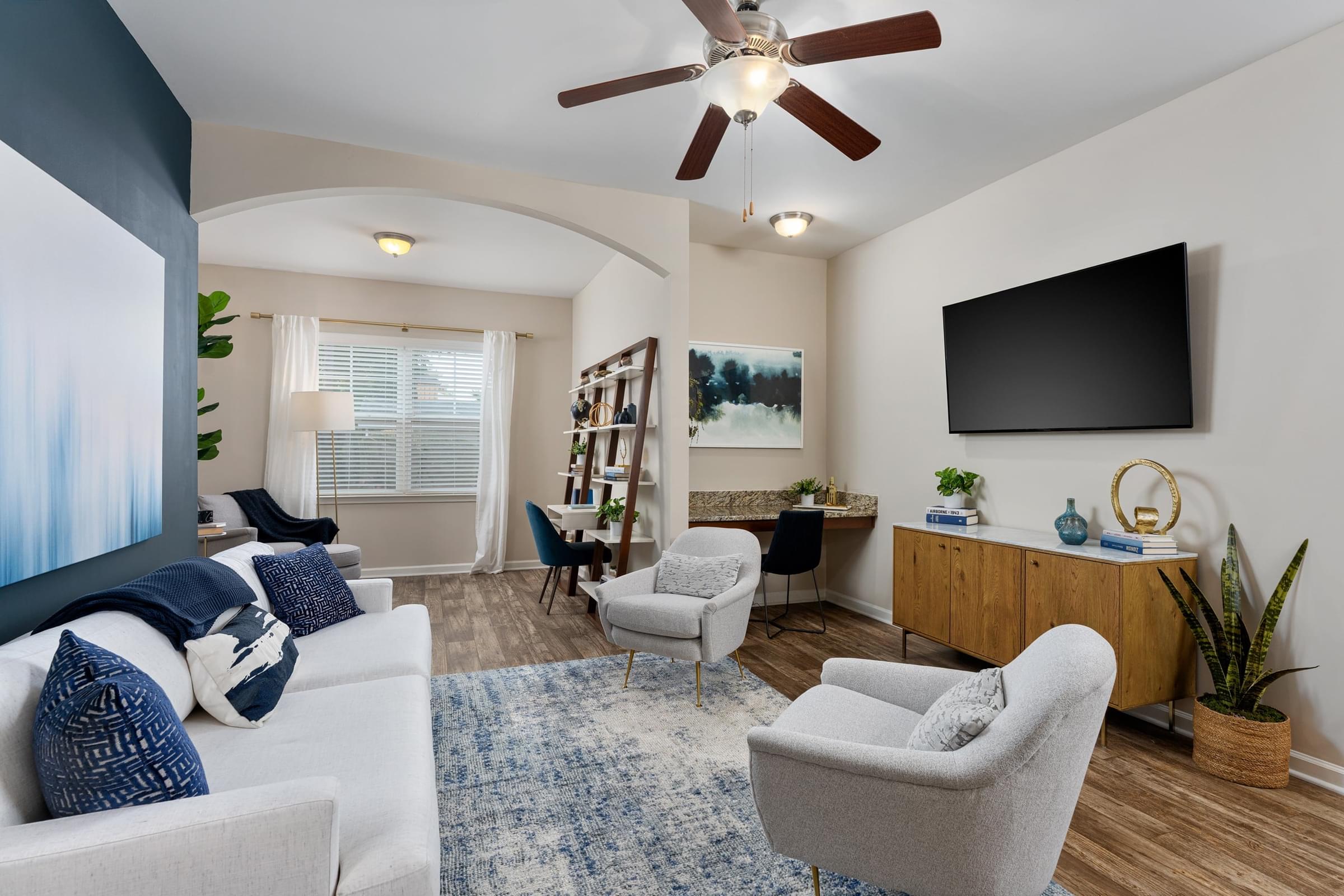 A living room with a white couch, a grey armchair, a wooden cabinet, a television, and a ceiling fan.