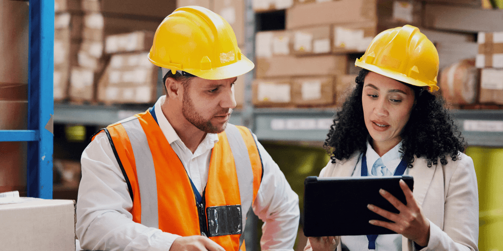 Two accounts receivable professionals in manufacturing review a tablet while wearing hard hats