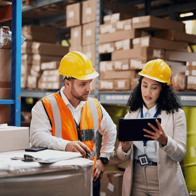 Two accounts receivable professionals in manufacturing review a tablet while wearing hard hats