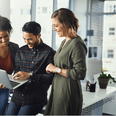 Three colleagues stand, smiling while looking at a laptop held by the middle person
