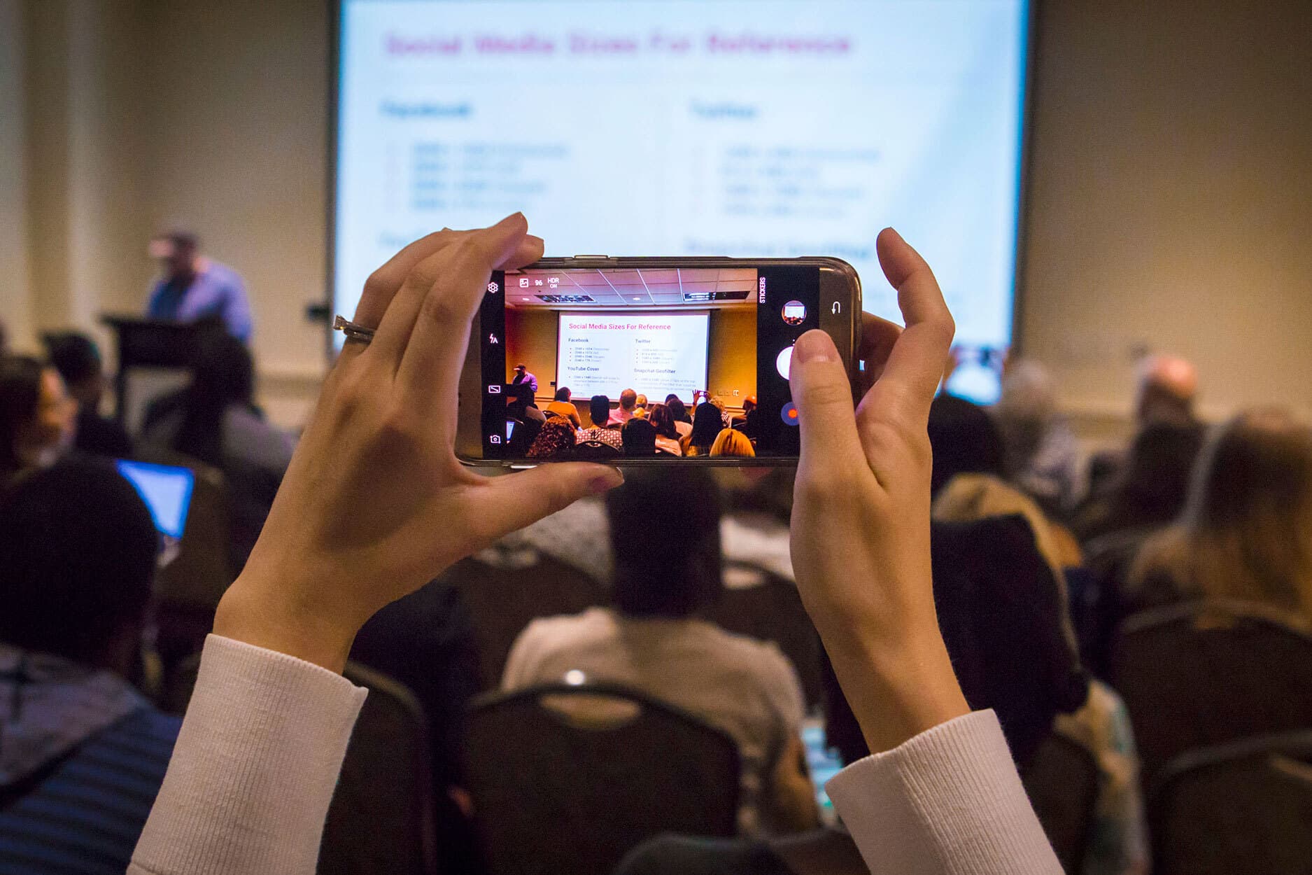 hands hold up a smartphone open to the camera app. the phone is pointed at the conferences main projection screen displaying a slide titled: Social media sizes for reference