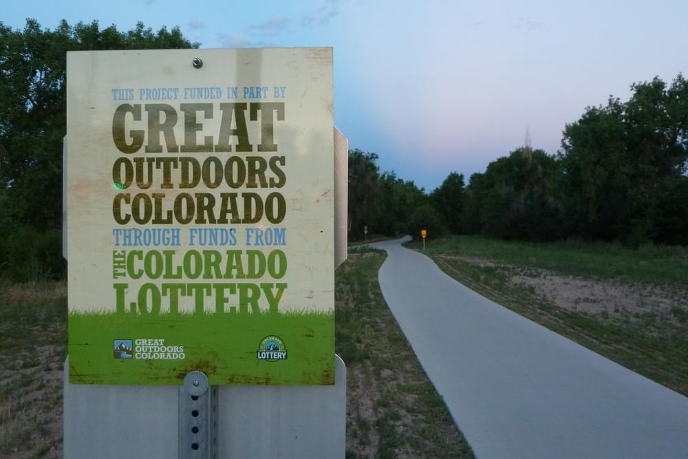 A newer style of sign boldly proclaiming the Colorado Lottery funding source on Cherry Creek Trail in Denver, Colorado