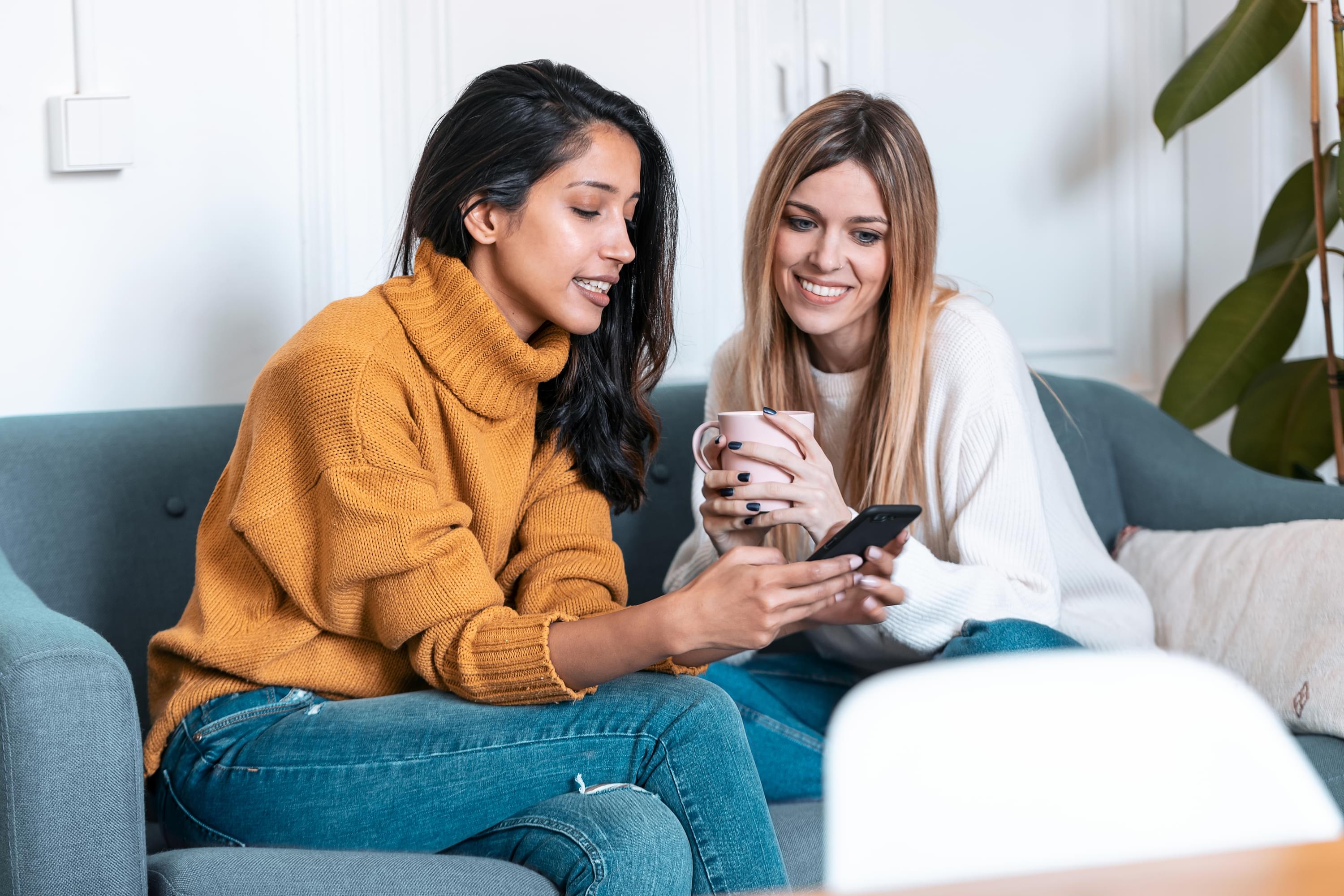 Shot two pretty young women using mobile phone while drinking coffee sofa home