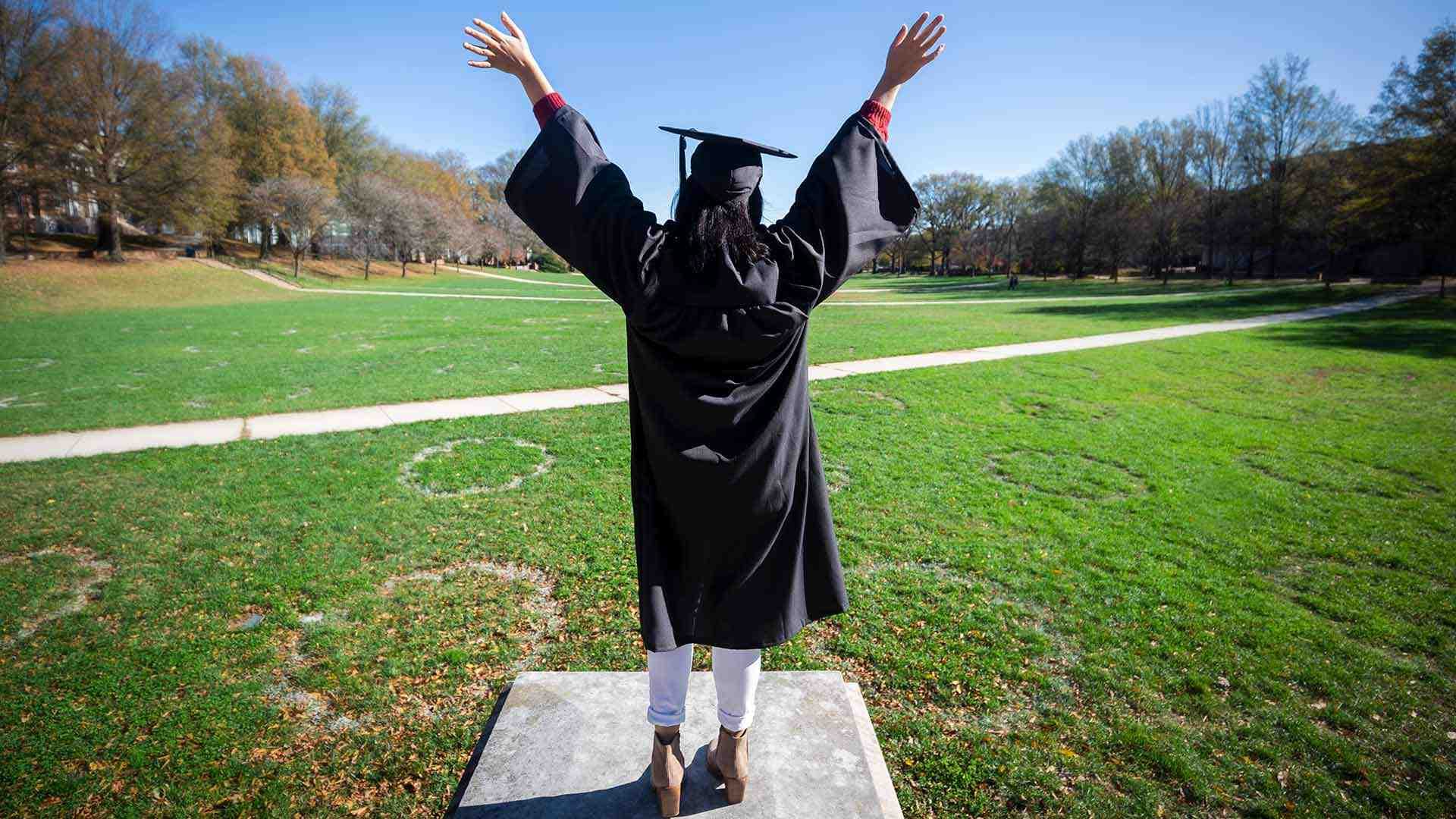 Student faced away from camera dressed in black cap and gown with arms lifted up in celebration against a green background of grass