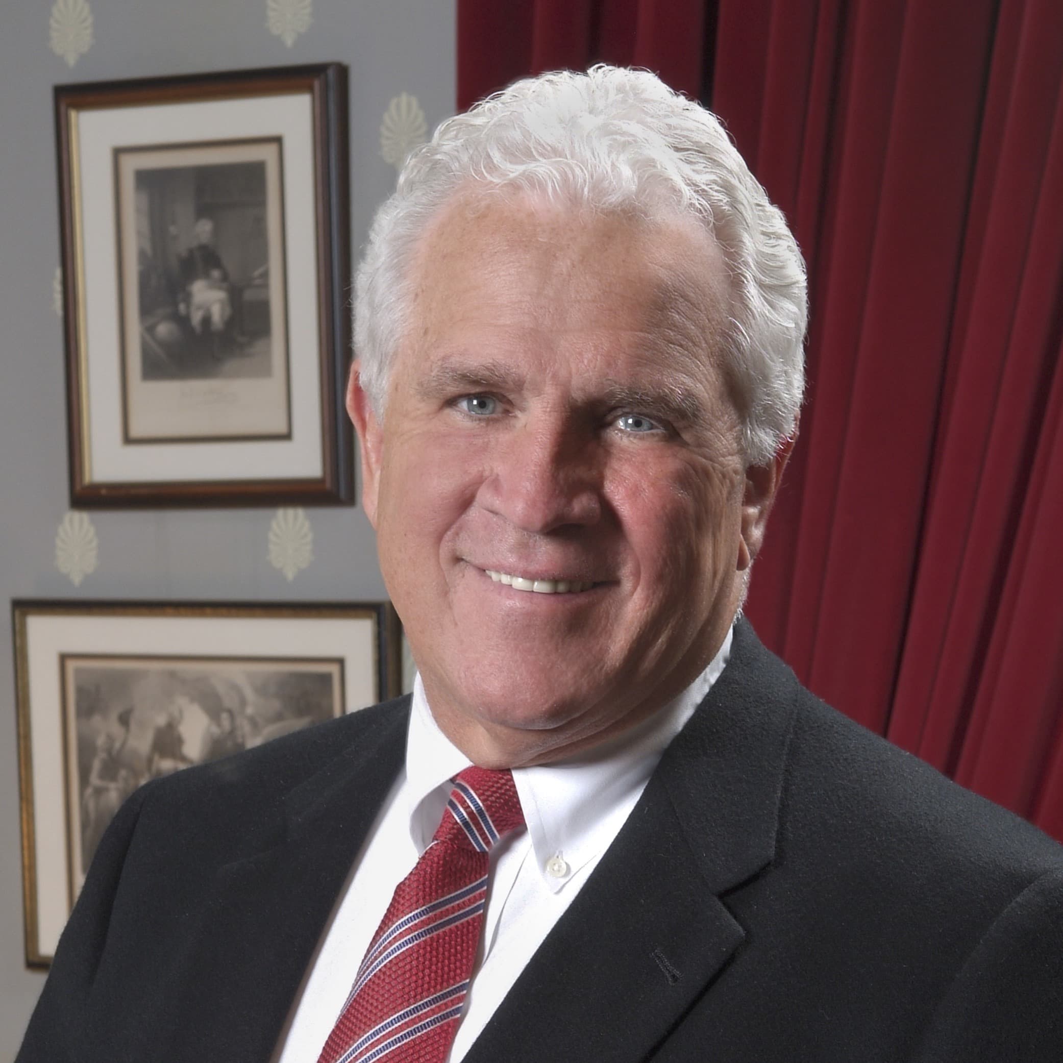 Full color headshot of Senate President Thomas V. "Mike" Miller, Jr. smiling wearing a black blazer, white dress shirt, and red striped tie against an office background. 