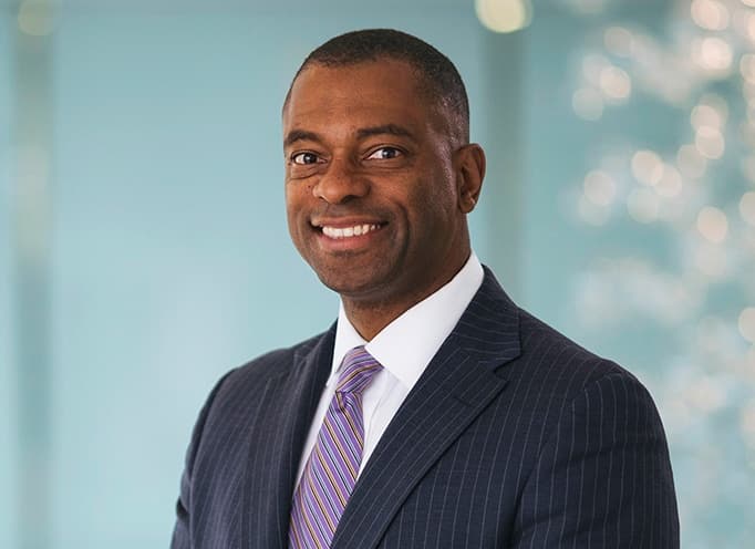 Full color headshot of Craig Thompson smiling wearing a navy pinstripe suit, white dress shirt, and striped tie against a light turquoise background.