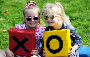Two little girls wearing sunglasses. They are smiling. The one on the left holds a red tile with a black X on it. The one on the right holds a yellow tile with a black O on it.