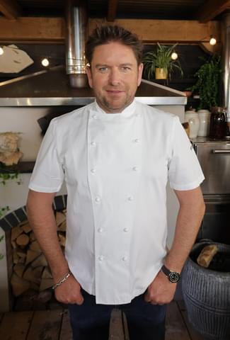 James Martin wearing chef whites, standing in front of a pizza oven.