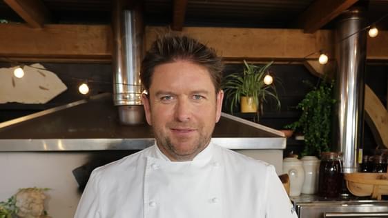 James Martin wearing chef whites, standing in front of a pizza oven.