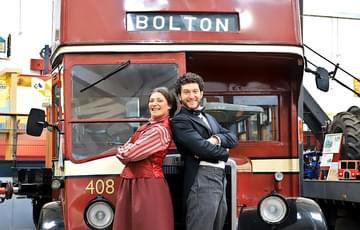 A woman and a man stand back-to-back with their arms crossed. Both dress in period clothing and stood in front of a vintage double-decker bus