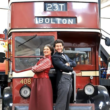 A woman and a man stand back-to-back with their arms crossed. Both dress in period clothing and stood in front of a vintage double-decker bus