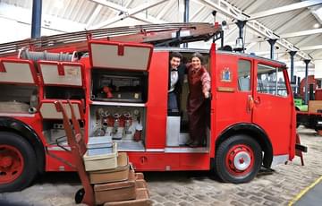 A man and woman both dressed in period clothing. They are stood in the carriage of an old vintage fire engine.