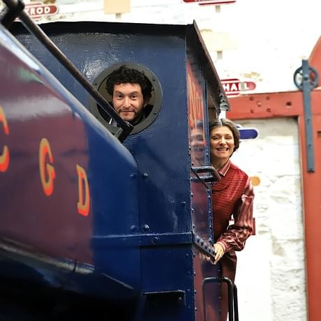 A man and a woman on a vintage steam train. The man is looking through a port hole and the woman is hanging out the side of the drivers carriage.
