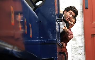 A man and a woman both dressed in period clothing. They are hanging out the carriage of a vintage steam train, the woman is pointing straight ahead.