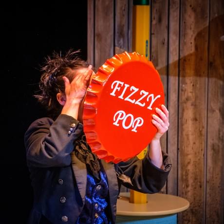 A person wears a long dark suit and holds a comically large bottle cap in front of their face. The bottle cap is read and reads 'Fizzy Pop' in white writing.