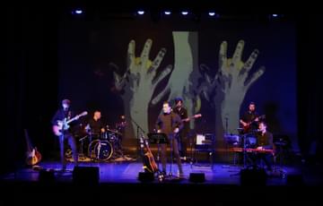 A band consisting of 6 people stand/sit on a lit stage. All playing different instruments such as: Guitar, Keyboard, Drums, Bass and one person looks like they will sing. The back wall has some large hands projected on to it.