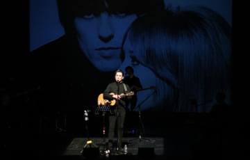 One person stands on stage in a spotlight, they are holding a guitar and singing. Two faces are projected onto the back wall of the stage.