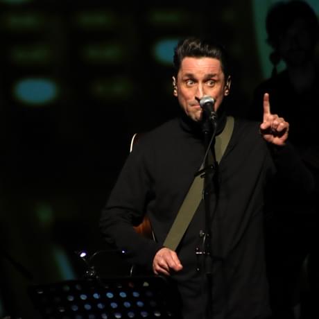 A person wearing all black, stands behind a microphone. They are holding one finger up on their left hand.