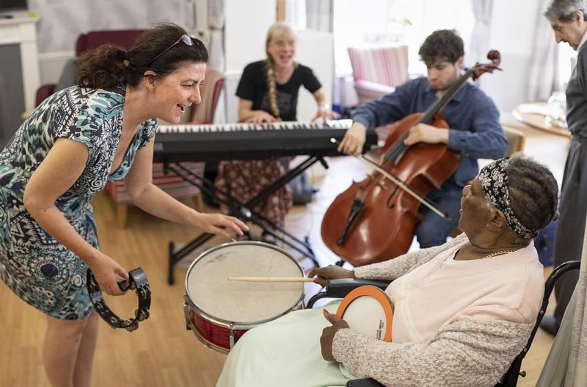 A woman with a tambourine gestures to an elderly lady playing a drum.