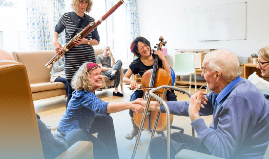 Musicians in a care setting. A woman smiles and shakes an older man's hand as other play instruments in front of them