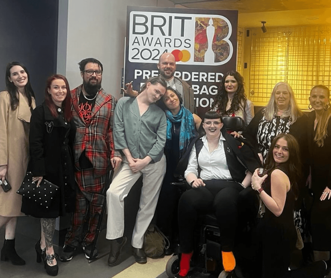 A group of people stand infront of a Brit Awards sign, smiling to the camera