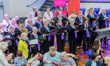 Image shows a choir of older women singing with a group of percussionists from a care home in front of them. They are taking part and performing in a community concert.