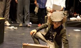 A young boy in a hat putting papers into a briefcase, with performers behind.