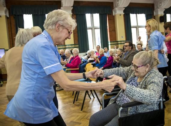 A care home staff member dancing with a seated elderly lady in a wheelchair.