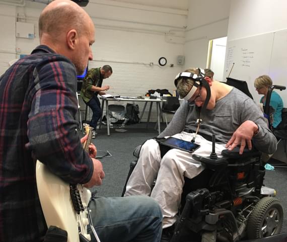A man playing a guitar with a man in a wheelchair using a pointer device attached to a headset.