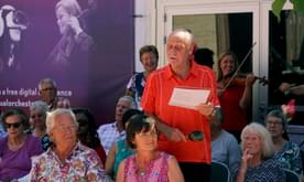 An older man stands in front of some musicians and sings from a sheet of paper.