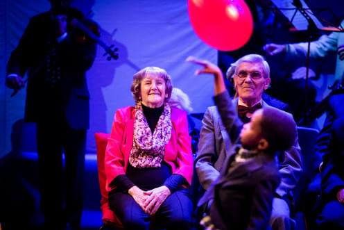 An older man and woman seated watching a young boy playing with a red balloon.