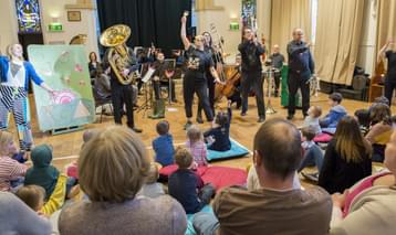 Musicians with brass instruments standing in front of a small orchestra, watched by an audience of adults and children.