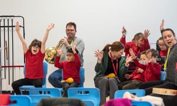 Children with their hands in their, sat next to musicians playing instruments