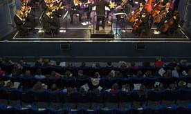 A audience of school children sat in theatre seating watching an orchestra perform on stage.