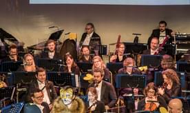 Two young producers dressed as a bee and lion present in front of an orchestra. A screen behind shows a musical score.