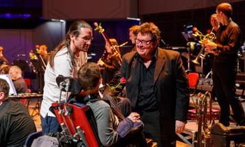 A young man in a wheelchair and his carer talk to a musician from the orchestra.
