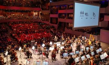 A full orchestra on stage performing to an audience of hundreds of school children, with a big screen overhead.