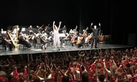 A presenter on stage in front of the orchestra waving to a large audience of children.
