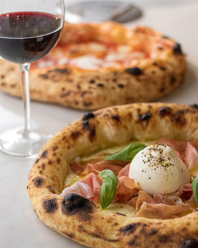 Two pizzas on a table next to a glass of wine.