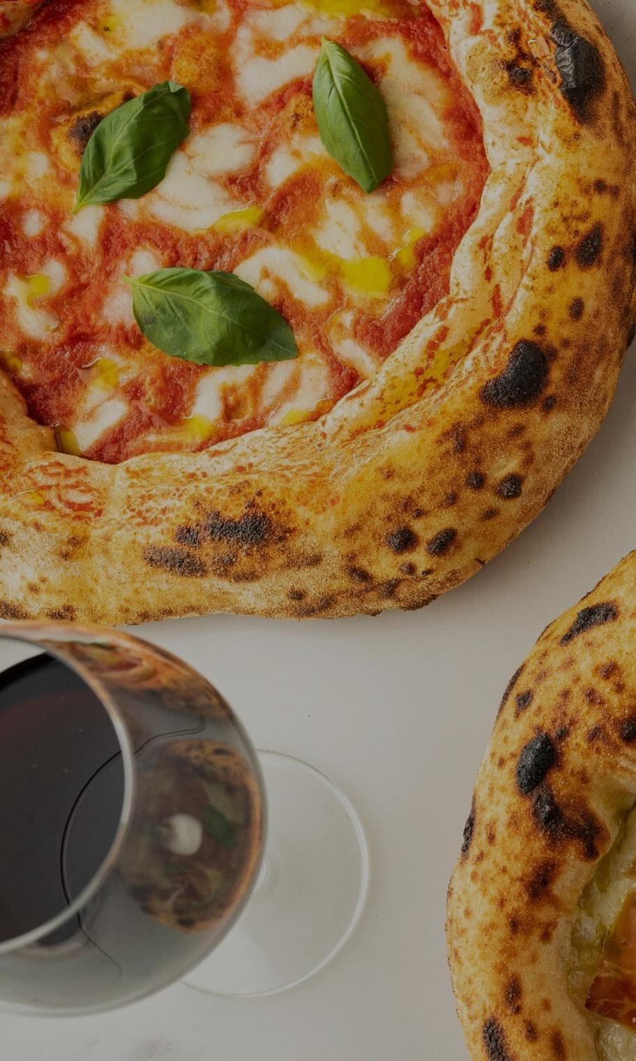Two pizzas on a table next to a glass of wine.